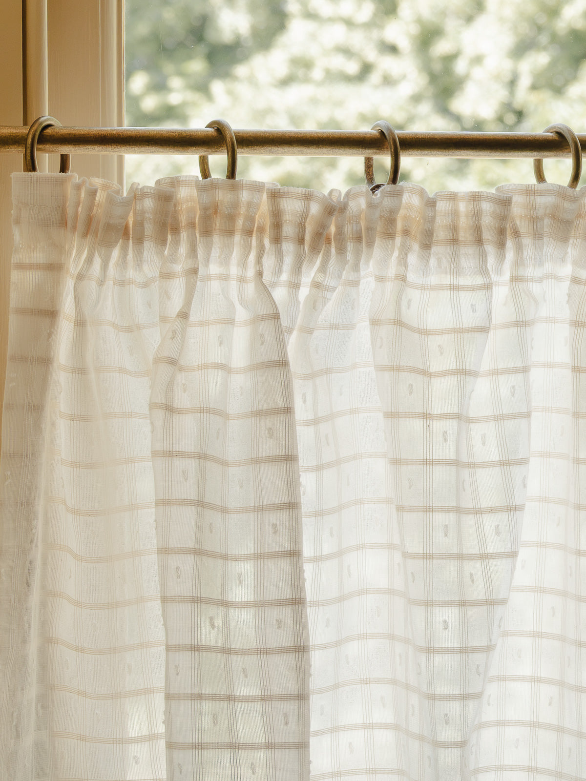 Close-up pleated café curtain in sheer Alda cotton on brass rings and pole in a bright hallway 