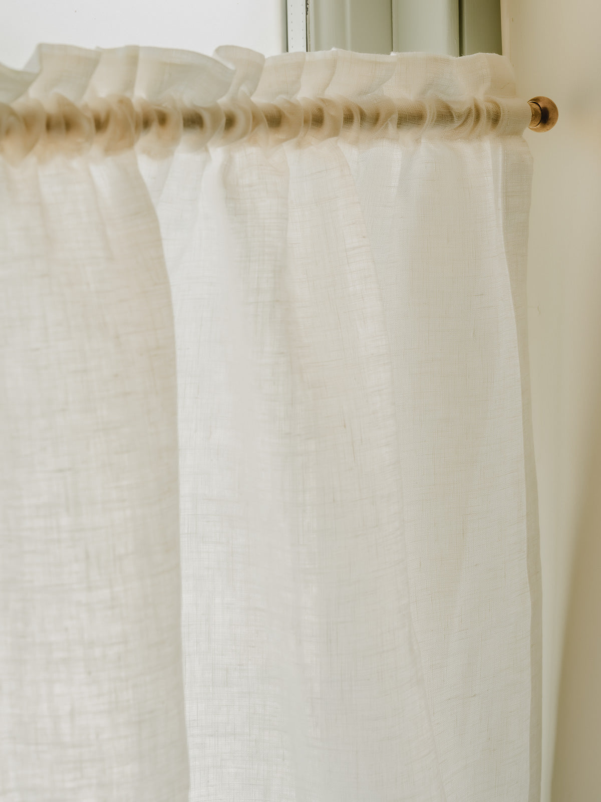 A close-up of a white sheer curtain panel hanging from a gold  rod, set against a soft beige wall and window frame.