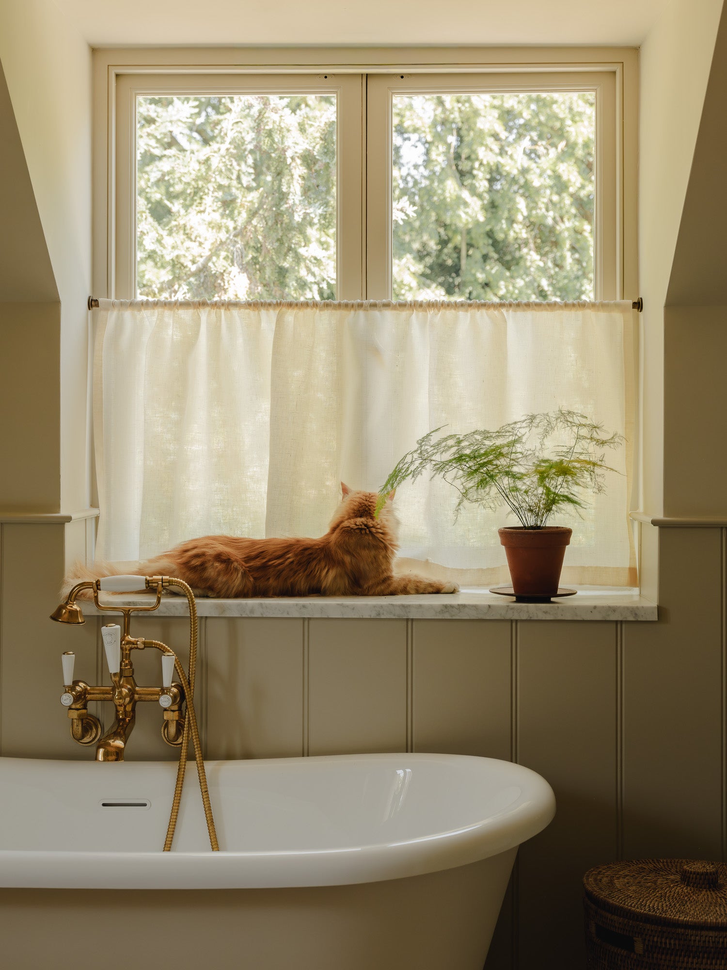 Rod pocket café curtain in cream Sera linen on a brass pole above a roll-top bath at Baywell Studio, ginger cat on the sill