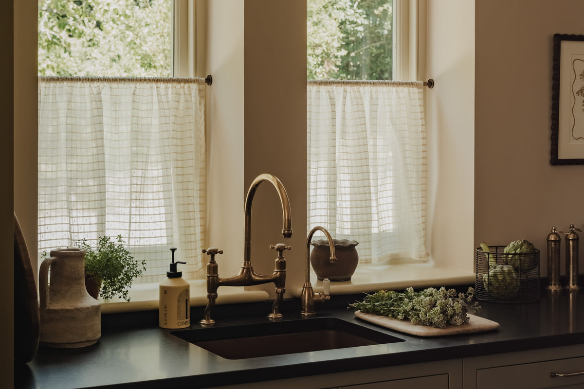Rod pocket café curtains in cream Alda filtering soft daylight into a Cotswold kitchen, on brass poles behind a copper sink