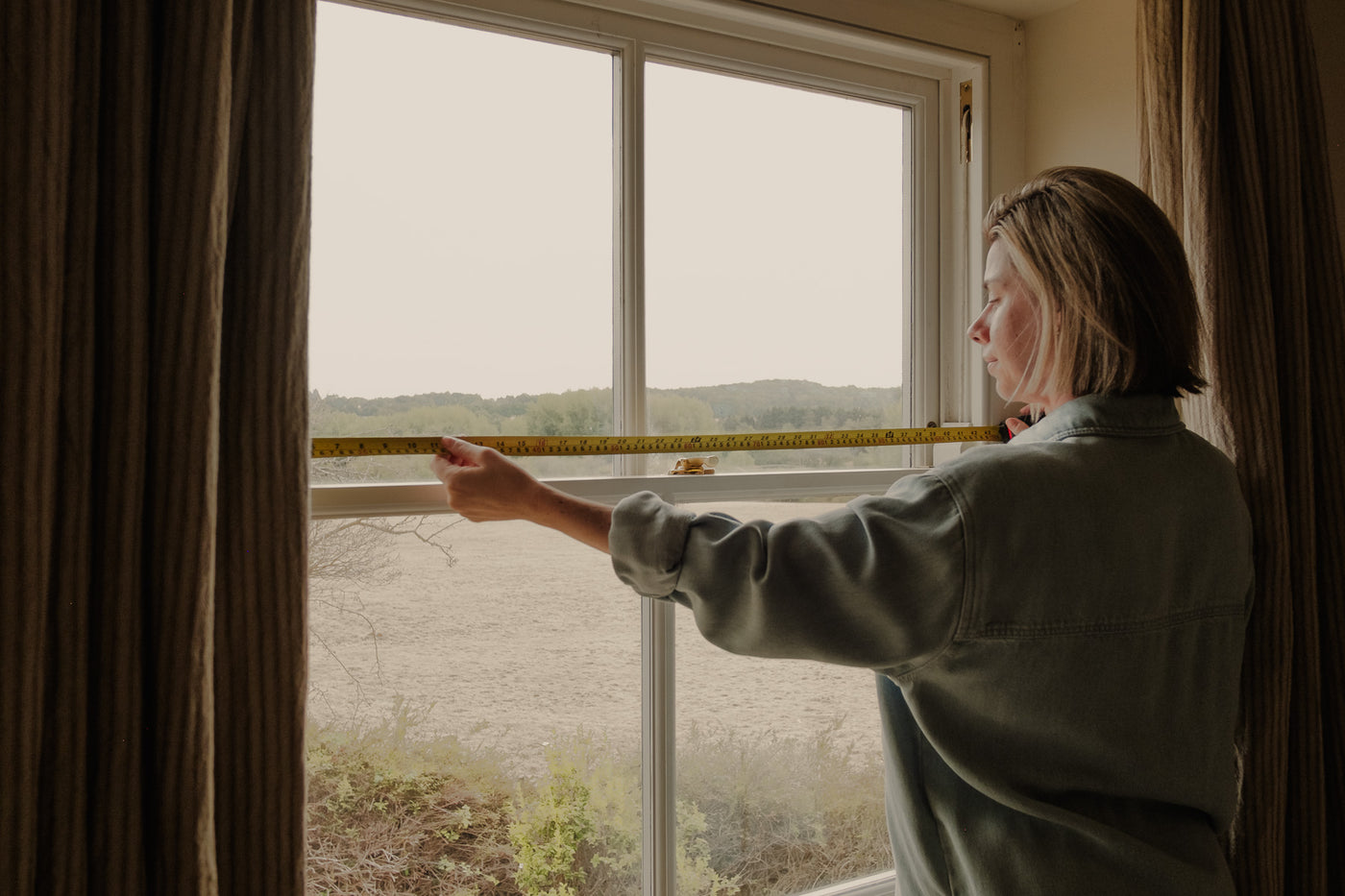 A woman stands by a large window, measuring window for curtains