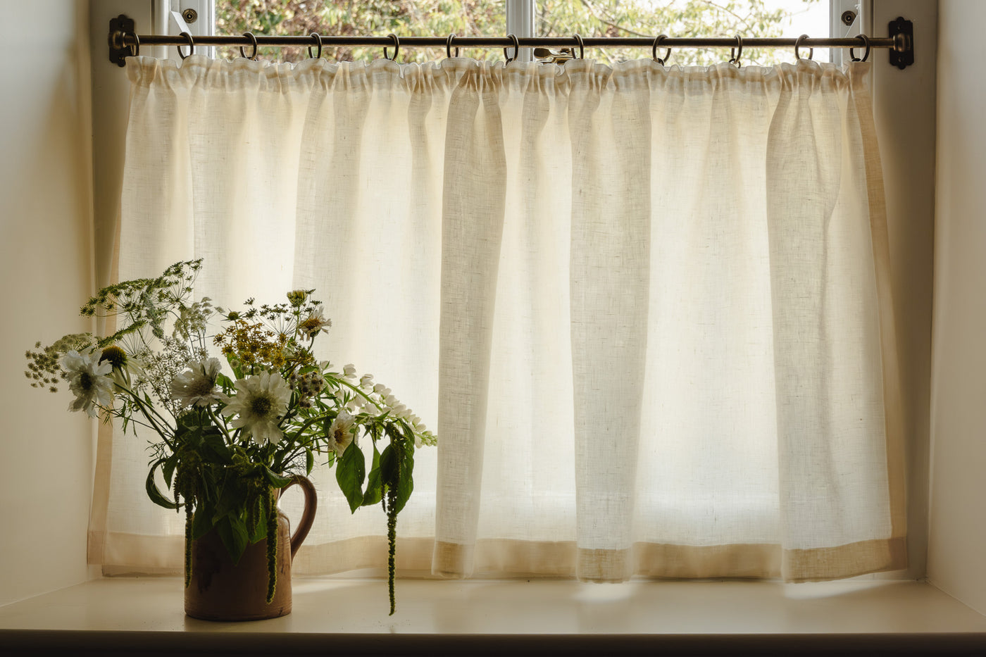 Cream linen cafe curtains hanging on a brass rod, styled with a vase of flowers.